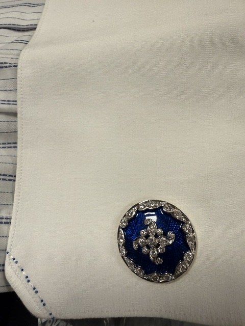 A white shirt cuff fastened with a round Gem Palace cufflink, blue guilloche enamel with a diamond swirl and diamond-set border
