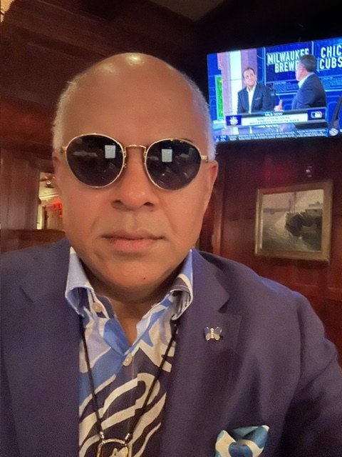 Sri Ramaswami in a wood-panelled bar, blue blazer and butterfly pin, WSY Edit